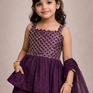 Op de afbeelding: Een jong meisje in een paarse outfit met gouden accenten. De top heeft een geschulpte halslijn en een gouden patroon. De outfit bevat een bijpassende broek en een doorschijnende sjaal. Gouden armbanden en een haaraccessoire maken de look compleet.