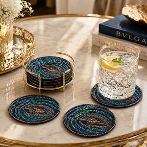 May include: A set of beaded coasters in shades of teal, blue, and gold, arranged on a marble-like surface. The coasters feature a leaf design and are held in a gold-colored metal holder. A glass of water with a lemon slice sits on one of the coasters.