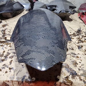 May include: A close-up of a black carbon fiber motorcycle part. The part is laid out on a light brown surface with dark brown stains.