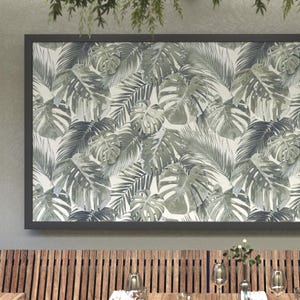 May include: A framed artwork with a tropical leaf design in grey and white. The image displays large monstera leaves and other foliage, creating a botanical pattern. The dark grey frame surrounds the artwork, which is displayed above a wooden slat wall, creating a natural aesthetic.