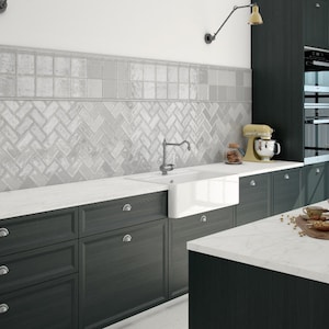 May include: A contemporary kitchen with dark grey cabinets and white worktops. The backsplash showcases a light grey herringbone tile design. A white farmhouse sink and a yellow stand mixer are present.