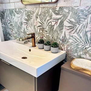 May include: A contemporary bathroom vanity featuring a white sink, copper tap, and three small potted succulents. The backsplash showcases a tropical leaf design in green and pink hues. A wooden tray rests on a matching cabinet.