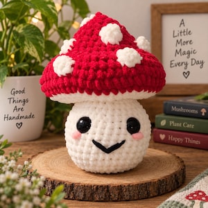 May include: A crocheted mushroom plush toy with a bright red cap featuring white spots and a white stem. The stem has a round face with large black safety eyes, pink stitched cheeks, and a black V-shaped mouth. The texture is soft and yarn-based.