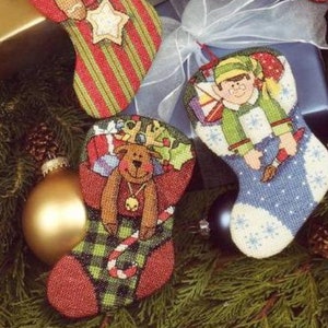 May include: Six colourful Christmas stocking ornaments with different holiday themes, including a gingerbread person, Santa Claus, an angel, an elf, a snowman, and a reindeer. The ornaments are displayed on a green pine branch with gold and blue gift boxes.