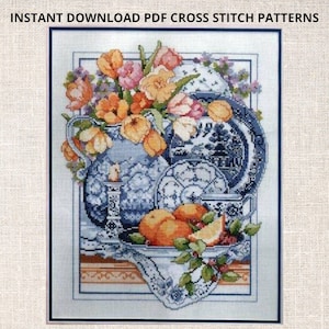Puede incluir: Patrón de punto de cruz con un jarrón de porcelana azul y blanco lleno de tulipanes naranjas y rosas, rodeado de platos y cuencos de porcelana azul y blanco, y un cuenco de naranjas y limones sobre un mantel de encaje blanco.