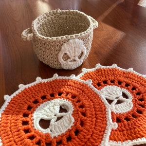 May include: Two orange crocheted coasters with white skull designs and a beige crocheted bowl with a white skull design.