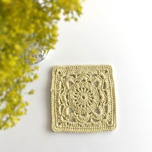 May include: A crocheted square coaster in a pale yellow color. The coaster features a delicate, intricate design with a central circle surrounded by smaller circles and loops.