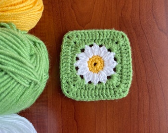 May include: A green crochet square with a white daisy in the centre. The daisy has a yellow centre and white petals. The square is surrounded by balls of yarn in yellow, green, and white.