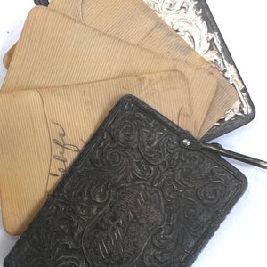 May include: A vintage black metal photo album with an ornate design. The album is closed and has a stack of old, brown paper envelopes inside.