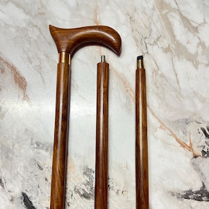 Puede incluir: Un bastón de madera con un mango curvo y un acabado marrón oscuro. El bastón está en tres piezas desmontables, con detalles metálicos color latón en las uniones. El bastón mide aproximadamente 91 cm de largo cuando está montado.