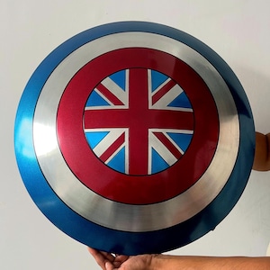 Puede incluir: Un escudo redondo con un borde azul, una banda plateada y un centro rojo con un diseño de la Union Jack. El escudo se muestra sobre un fondo blanco.