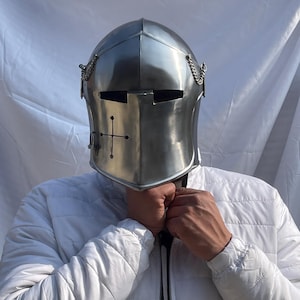 Peut inclure: Casque de chevalier argent&eacute; poli avec un embl&egrave;me de croix &agrave; l'avant. Le casque comporte des fentes pour les yeux et des d&eacute;tails de cha&icirc;ne. L'image montre le casque sur une personne portant une veste blanche.