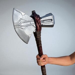 May include: A silver and gray battle axe with a dark brown handle. The axe head has a rectangular shape with a silver blade and a gray hammerhead. The handle is wrapped in brown leather.
