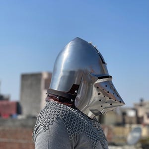 May include: A shiny, silver-coloured medieval helmet with a pointed top and a visor with small holes. The helmet is worn over a chainmail coif and a grey shirt. A brown leather strap is visible at the base of the helmet.