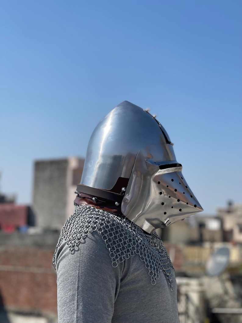 May include: A shiny, silver-coloured medieval helmet with a pointed top and a visor with small holes. The helmet is worn over a chainmail coif and a grey shirt. A brown leather strap is visible at the base of the helmet.