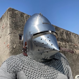May include: A shiny, silver-coloured medieval helmet with a pointed visor and small ventilation holes. The helmet is worn with a chainmail coif and leather straps. The image is taken outdoors against a clear blue sky.