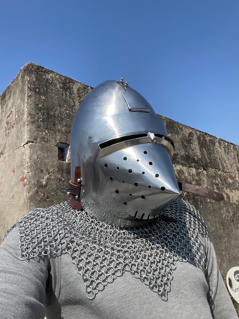 May include: A shiny, silver-coloured medieval helmet with a pointed visor and small ventilation holes. The helmet is worn with a chainmail coif and leather straps. The image is taken outdoors against a clear blue sky.