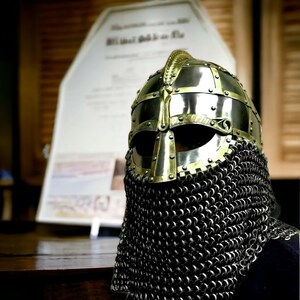 Puede incluir: Un casco medieval de metal dorado con protecci&oacute;n para el cuello de cota de malla. El casco tiene una forma redondeada, una barra nasal y aberturas para los ojos. La cota de malla est&aacute; hecha de peque&ntilde;os anillos met&aacute;licos entrelazados.