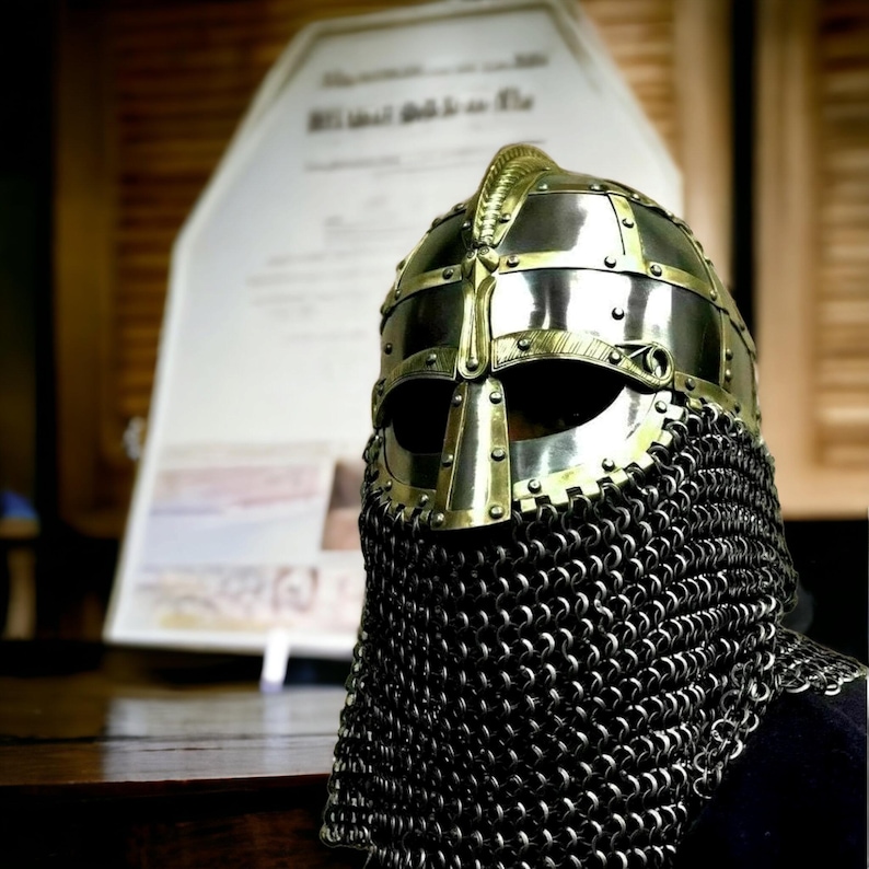 Puede incluir: Un casco medieval de metal dorado con protecci&oacute;n para el cuello de cota de malla. El casco tiene una forma redondeada, una barra nasal y aberturas para los ojos. La cota de malla est&aacute; hecha de peque&ntilde;os anillos met&aacute;licos entrelazados.