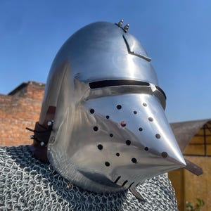 May include: A shiny, silver-coloured medieval helmet with a pointed visor and many small ventilation holes. The helmet has a rounded top and is secured with leather straps. A chainmail shirt is visible below the helmet.