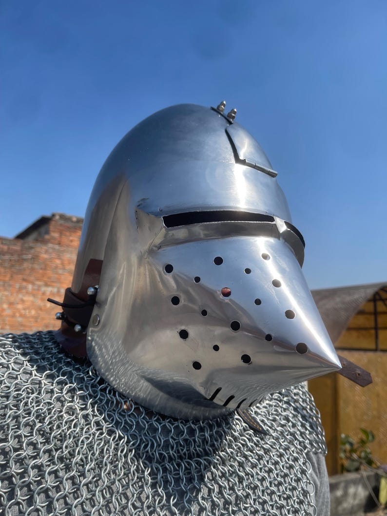 May include: A shiny, silver-coloured medieval helmet with a pointed visor and many small ventilation holes. The helmet has a rounded top and is secured with leather straps. A chainmail shirt is visible below the helmet.