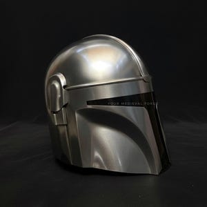 May include: A silver-colored Mandalorian helmet with a black visor. The helmet has a smooth, metallic finish and is designed with angular shapes. The text "YOUR MEDIEVAL FORCE" is visible on the visor.