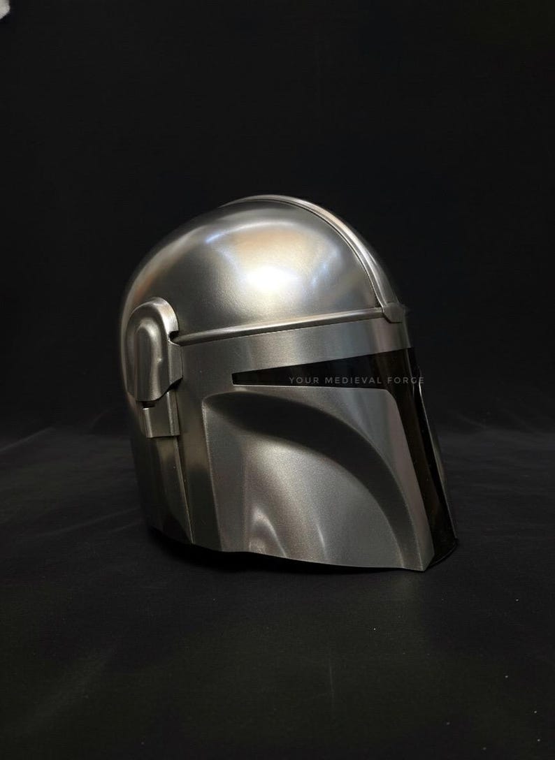 May include: A silver-colored Mandalorian helmet with a black visor. The helmet has a smooth, metallic finish and is designed with angular shapes. The text "YOUR MEDIEVAL FORCE" is visible on the visor.