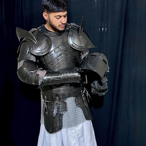 K&ouml;nnte beinhalten: Eine komplette R&uuml;stung aus schwarzem Metall, einschlie&szlig;lich Helm, Schulterschutz und Brustplatte. Die R&uuml;stung wird &uuml;ber Kettenhemden und einem wei&szlig;en Kleidungsst&uuml;ck getragen. Die Person h&auml;lt den Helm.