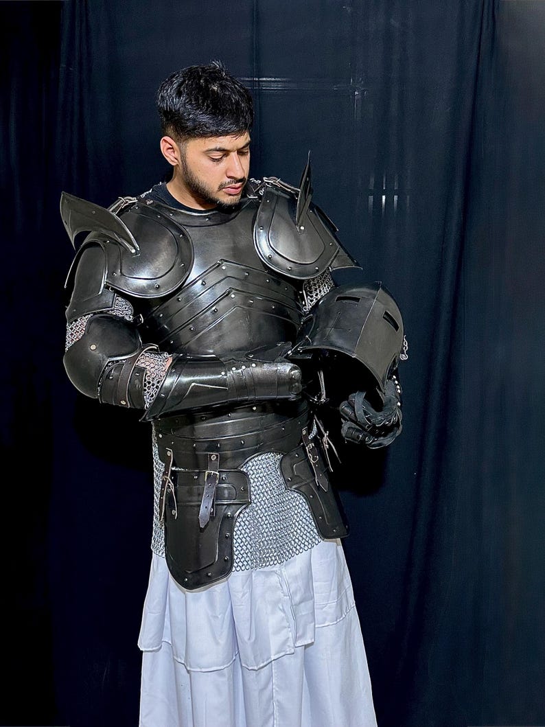 K&ouml;nnte beinhalten: Eine komplette R&uuml;stung aus schwarzem Metall, einschlie&szlig;lich Helm, Schulterschutz und Brustplatte. Die R&uuml;stung wird &uuml;ber Kettenhemden und einem wei&szlig;en Kleidungsst&uuml;ck getragen. Die Person h&auml;lt den Helm.