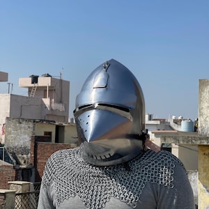 May include: A shiny, silver medieval helmet with a pointed visor and rivet details. The helmet is worn over a grey long-sleeved top and chainmail armour. The background shows a clear blue sky and buildings.