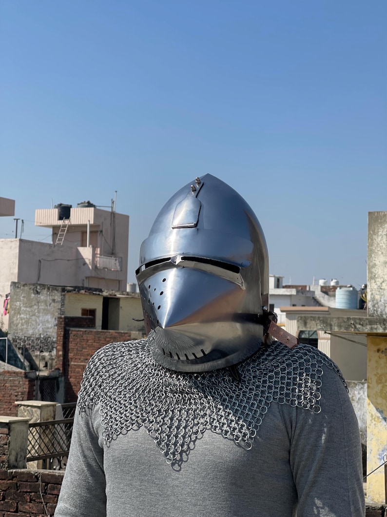 May include: A shiny, silver medieval helmet with a pointed visor and rivet details. The helmet is worn over a grey long-sleeved top and chainmail armour. The background shows a clear blue sky and buildings.