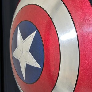May include: A red, white, and blue shield with a silver rim and a white star in the center. The shield has a metallic finish and a circular design. The shield is a replica of Captain America's shield.