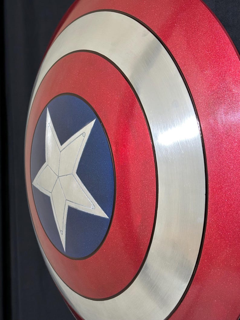 May include: A red, white, and blue shield with a silver rim and a white star in the center. The shield has a metallic finish and a circular design. The shield is a replica of Captain America's shield.