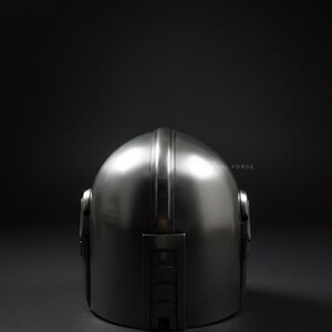 May include: A metallic silver helmet with a rounded top and a vertical ridge down the center. The helmet has a smooth, reflective surface and is set against a dark background. The helmet's design includes side features and a rectangular section at the back.