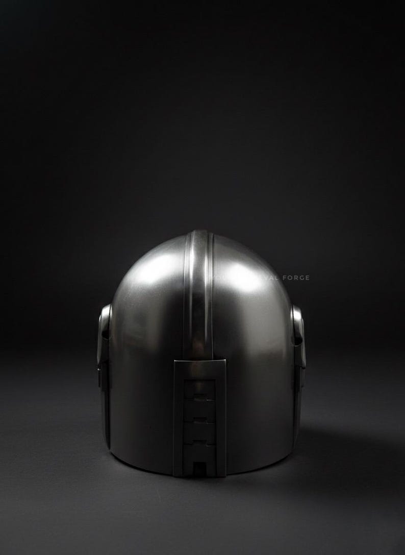 May include: A metallic silver helmet with a rounded top and a vertical ridge down the center. The helmet has a smooth, reflective surface and is set against a dark background. The helmet's design includes side features and a rectangular section at the back.