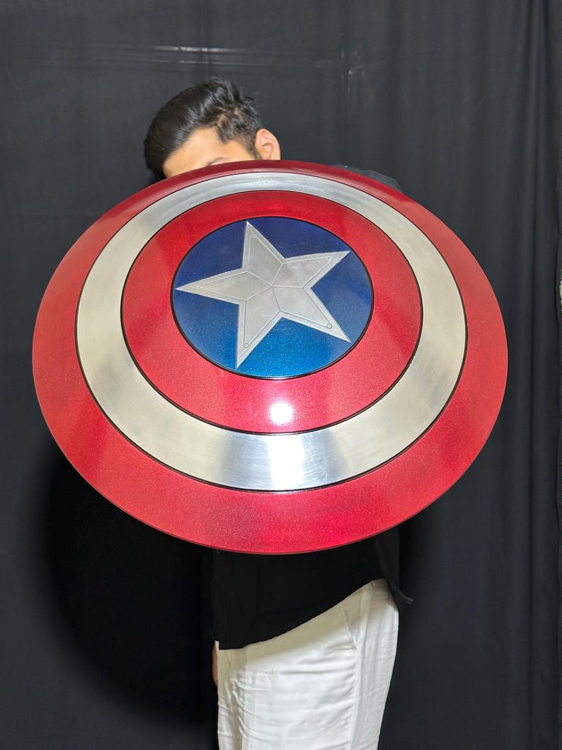 May include: A round shield with a red, silver, and blue color scheme. The shield features a central blue circle with a white star, surrounded by silver and red concentric rings. The shield is a replica of Captain America's shield.