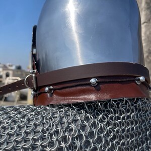 May include: A polished silver medieval helmet with a brown leather neck guard and a chainmail coif. The helmet has a rounded top and a dark leather strap. The chainmail is made of small, interlocking metal rings.