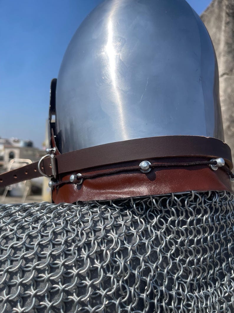 May include: A polished silver medieval helmet with a brown leather neck guard and a chainmail coif. The helmet has a rounded top and a dark leather strap. The chainmail is made of small, interlocking metal rings.