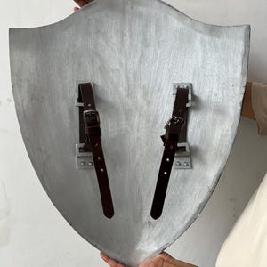 May include: A silver-colored shield with a curved top and pointed bottom. Two brown leather straps with buckles are attached to the back with metal brackets. The shield is held by two hands.