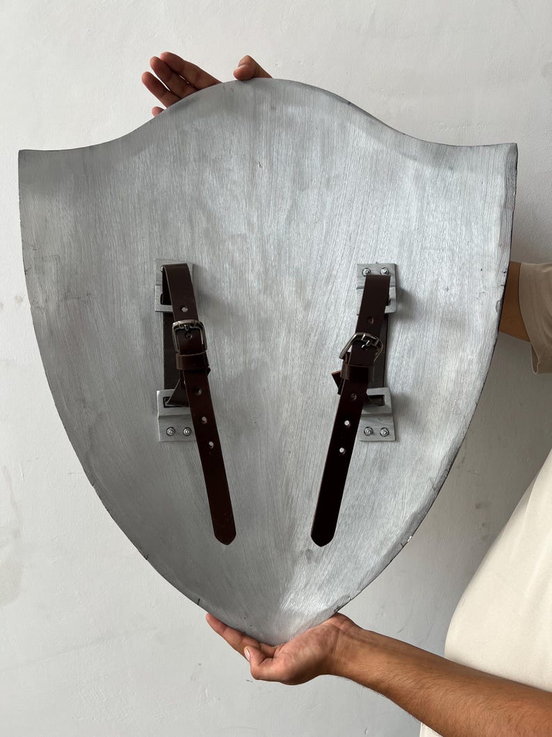 May include: A silver-colored shield with a curved top and pointed bottom. Two brown leather straps with buckles are attached to the back with metal brackets. The shield is held by two hands.
