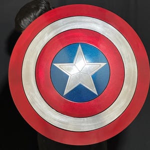 May include: A round shield with a red outer ring, a silver middle ring, and a blue center with a silver star. The shield has a metallic finish and is a replica of Captain America's shield.