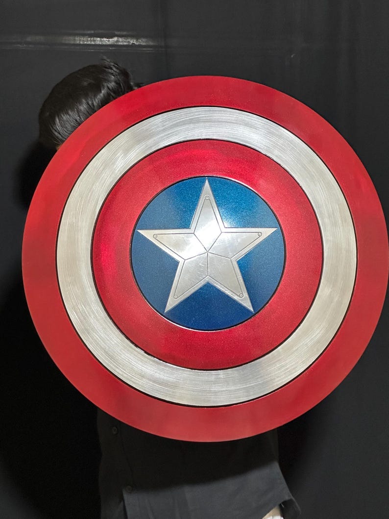 May include: A round shield with a red outer ring, a silver middle ring, and a blue center with a silver star. The shield has a metallic finish and is a replica of Captain America's shield.