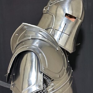 May include: A polished, silver-colored medieval knight's helmet and shoulder armor. The helmet features a cross design and eye slits. The shoulder armor is curved and riveted, with chainmail visible beneath. The overall design is reflective and detailed.