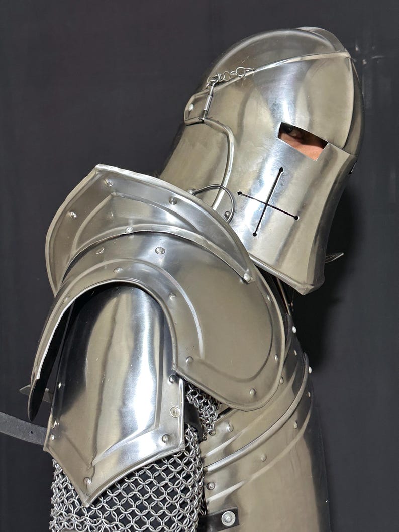 May include: A polished, silver-colored medieval knight's helmet and shoulder armor. The helmet features a cross design and eye slits. The shoulder armor is curved and riveted, with chainmail visible beneath. The overall design is reflective and detailed.