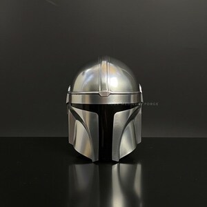 May include: A silver-colored Mandalorian helmet with a black visor. The helmet has a smooth, metallic finish and is designed with distinct angular features. The image is set against a dark background, highlighting the helmet's reflective surface.