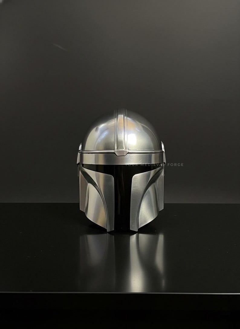 May include: A silver-colored Mandalorian helmet with a black visor. The helmet has a smooth, metallic finish and is designed with distinct angular features. The image is set against a dark background, highlighting the helmet's reflective surface.