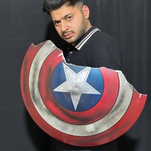 Op de afbeelding: Een Captain America schild prop met een distressed afwerking. Het schild heeft een blauwe centrale cirkel met een witte ster, omringd door rode en witte concentrische ringen. Het schild heeft een gevechtsversleten uiterlijk.
