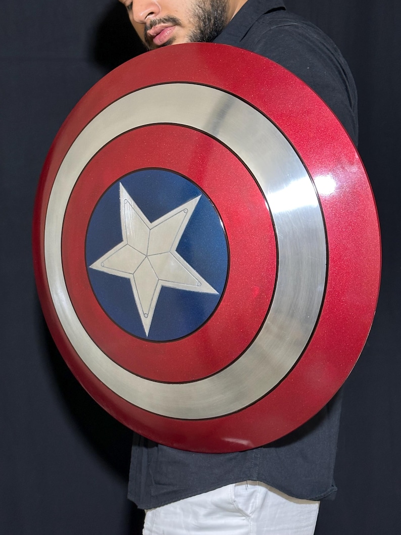 May include: A red, white, and blue shield with a star in the center. The shield has a circular design with a silver border and a blue circle in the middle. The shield is a prop or collectible.