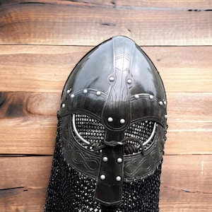 May include: A black Viking helmet with a riveted design and a chainmail neck guard. The helmet features intricate metalwork around the eye and mouth openings. The helmet is displayed against a wooden background.