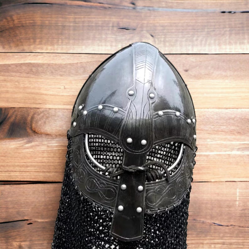 May include: A black Viking helmet with a riveted design and a chainmail neck guard. The helmet features intricate metalwork around the eye and mouth openings. The helmet is displayed against a wooden background.